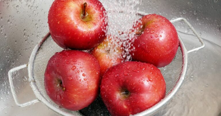 Closeup of red apples in colander washing under running water in sink.